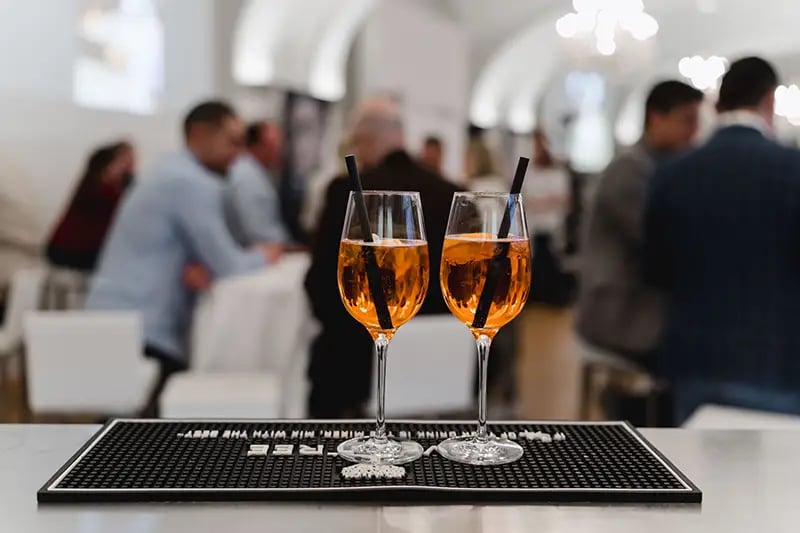 Auf dem Bild sieht man ein Tablett mit 2 Gläsern mit Aperol im Fokus, im Hintergrund sind Teilnehmer:innen sichtbar.